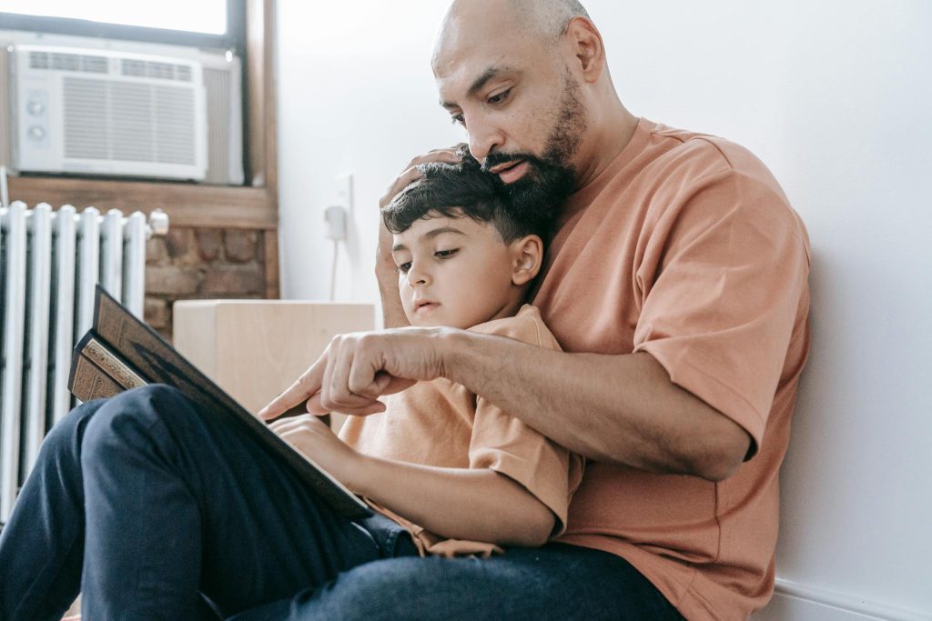A father in an orange shirt reads a book to his son while sitting indoors, fostering learning and bonding.